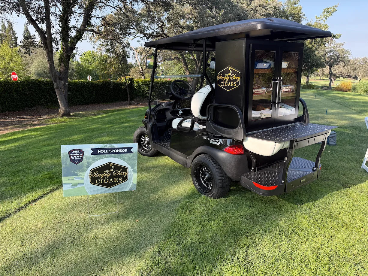 The black Simply Sexy Cigars mobile cigar cart with its glass humidor parked on a golf course beside a Hole Sponsor sign